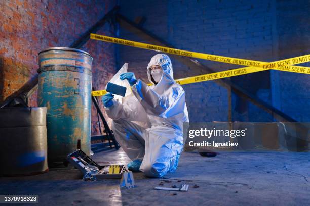 criminologist in a protective suit against the barricade tape holds a physical evidence in a plastic bag - forensisch stockfoto's en -beelden