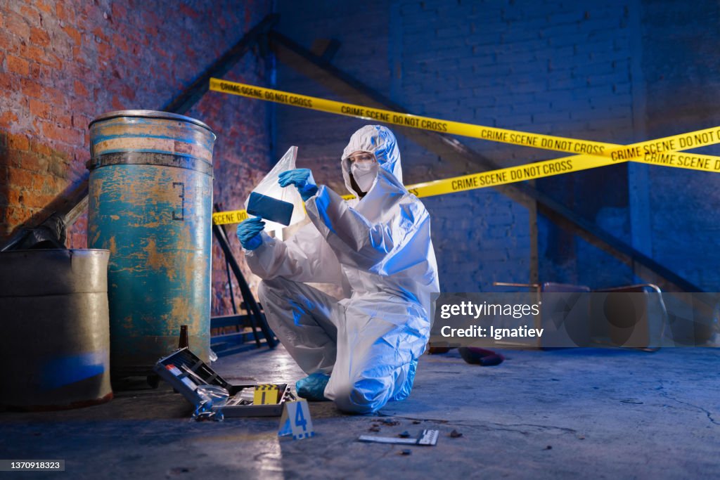 Criminologist in a protective suit against the barricade tape holds a physical evidence in a plastic bag