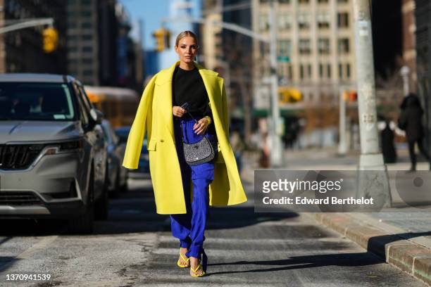 Leonie Hanne wears black sunglasses, a black and silver Prada pendant earring, a black high neck wool pullover, a yellow oversized long coat, high...