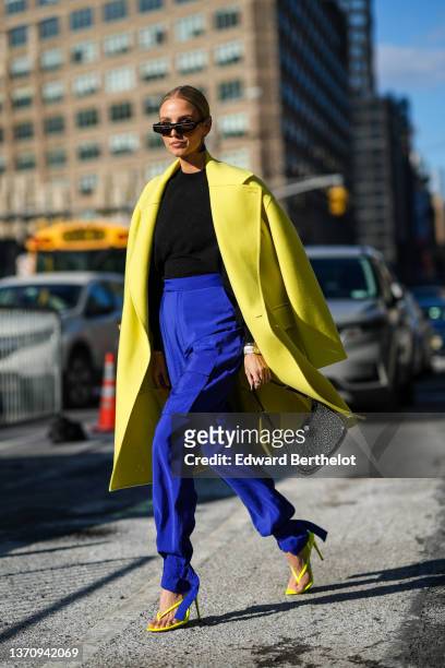 Leonie Hanne wears black sunglasses, a black and silver Prada pendant earring, a black high neck wool pullover, a yellow oversized long coat, high...