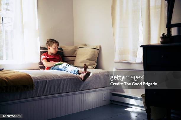 young boy reading book in bedroom at home - boy sitting on bed stock-fotos und bilder