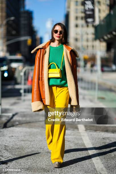 Guest wears black sunglasses, a green ribbed wool pullover, a camel shiny leather and beige sheep interior oversized aviator jacket, a yellow shiny...