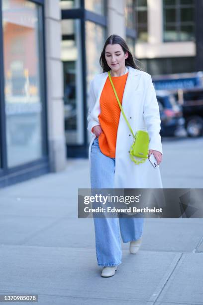 Guest wears a white t-shirt, a white long coat, a neon orange pullover, blue faded denim wide legs pants, a neon green crossbody bag with a large...