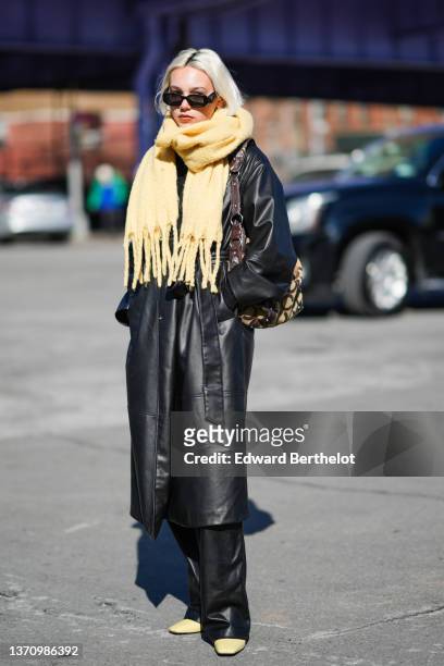 Guest wears black sunglasses from Prada, a pale yellow fringed wool scarf, a black shiny leather long coat, black shiny leather flared pants, a pale...