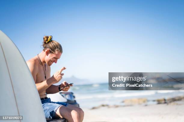 young male surfer with fingers crossed gambling online using phone at the beach - fingers crossed stock pictures, royalty-free photos & images