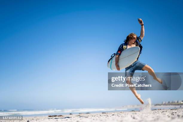 surfista extático saltando en el aire en la playa sosteniendo su teléfono - surfing the net fotografías e imágenes de stock