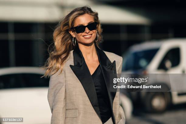 Xenia Adonts wears black sunglasses, large silver earrings, a black V-neck t-shirt, a beige checkered print pattern with black satin collar oversized...