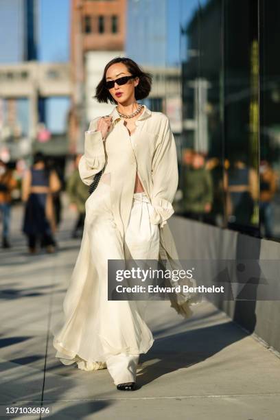 Chriselle Lim wears black sunglasses, gold earrings, a silver shiny pearls necklace, a white lace pattern V-neck cropped top, a beige long oversized...