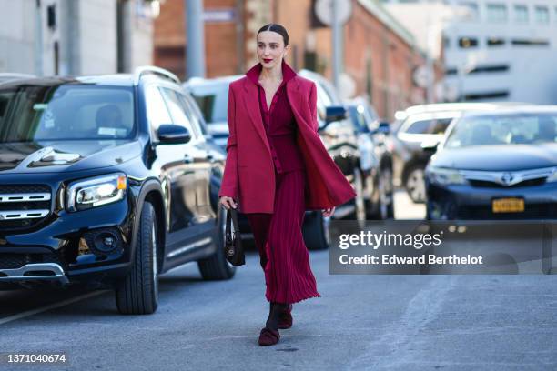 Mary Leest wears a burgundy pleated / accordion buttoned shirt, a matching burgundy pleated / accordion buttoned midi skirt, a black Re-edition 2000...