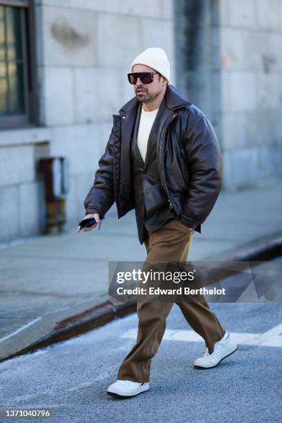 Alex Badia wears a white ribbed wool beanie, black sunglasses, a white pullover, a black shiny leather long blazer jacket, a black shiny leather...