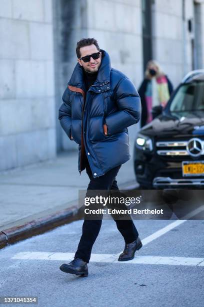 Derek Blasberg wears black sunglasses, a black turtleneck pullover, a black oversized puffer jacket, black denim jeans pants, black shiny leather...