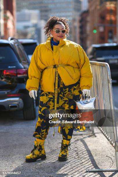 Greivy Lou wears beige / brown checkered print pattern sunglasses, gold earrings, a royal blue electric turtleneck t-shirt, a black with brown and...