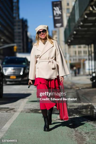 Guest wears a white braided wool beanie, black sunglasses, a beige winter coat, a red ruffled midi dress, a red shiny leather large handbag, black...