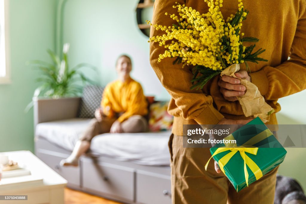 Young man about to surprise his girlfriend with a bouquet of mimosa flowers and a gift box