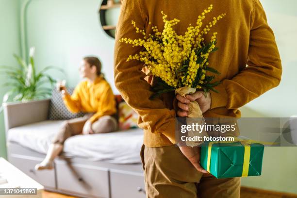 giovane premuroso con un mazzo di fiori di mimosa e una scatola regalo dietro la schiena - mimosa foto e immagini stock