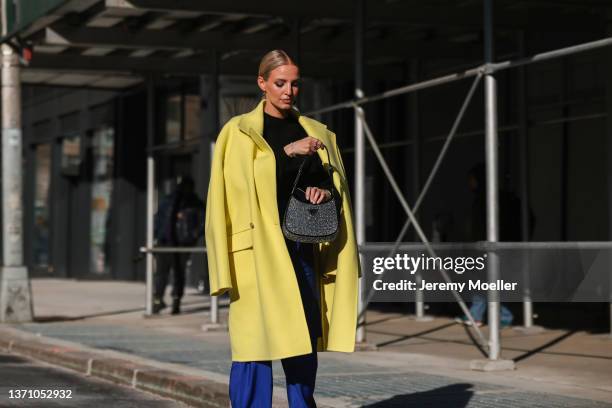 Leonie Hanne wears black sunglasses, a black and silver Prada pendant earring, a black high neck wool pullover, a yellow oversized long coat, high...