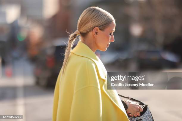 Leonie Hanne wears black sunglasses, a black and silver Prada pendant earring, a black high neck wool pullover, a yellow oversized long coat, high...