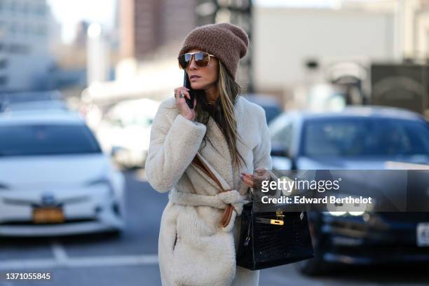 Guest wears a brown wool beanie, brown sunglasses, a white sheep long coat, a black shiny leather crocodile print pattern Kelly handbag from Hermes,...