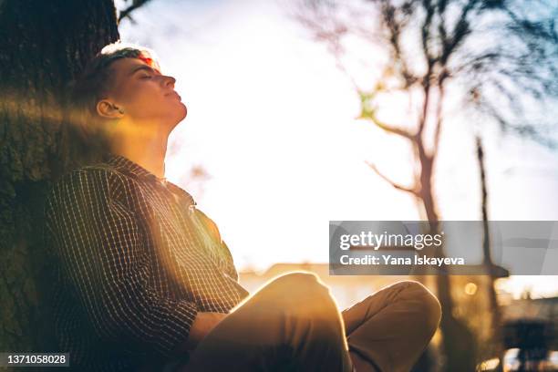 young handsome hipster is taking a rest under the tree, enjoying warm sunshine - benessere mentale foto e immagini stock