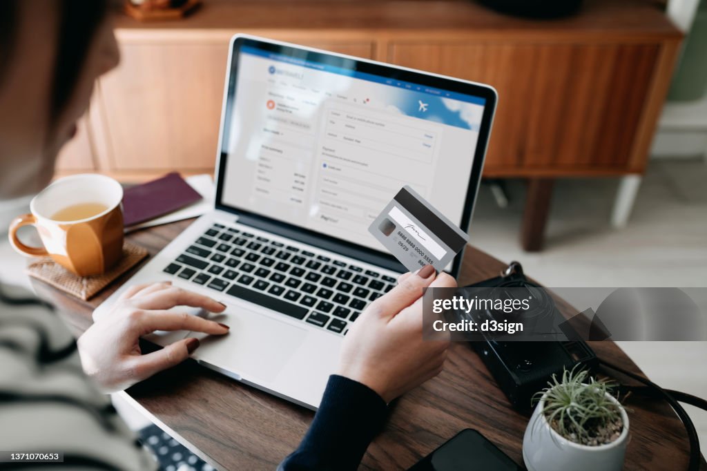 Over the shoulder view of young Asian woman shopping online for flight tickets on airline website with laptop, entering credit card details to make mobile payment at home. Camera and passport on the table. Travel planning. Booking a holiday online