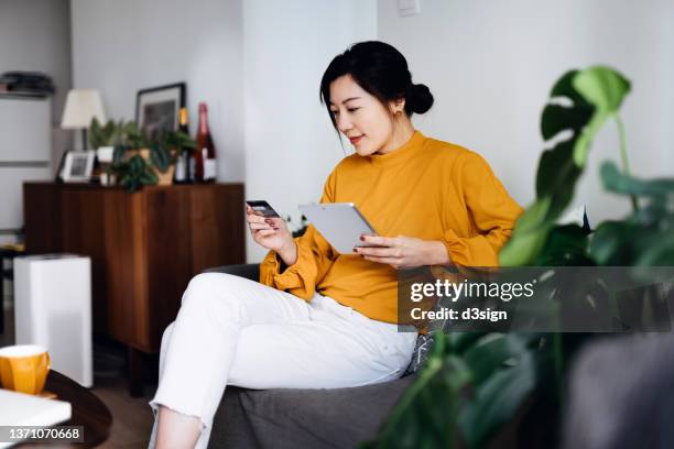 beautiful smiling young asian woman sitting on sofa in the living room, managing online banking, handling bank account and financial bills with digital tablet and credit card at home. convenience and easy banking. smart banking with technology - customer confidence stock pictures, royalty-free photos & images