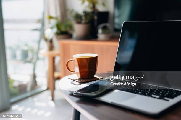 cropped shot of wooden coffee table with laptop, smartphone and a cup of tea in the living room at home by the window against beautiful sunlight - coffee cups table stockfoto's en -beelden