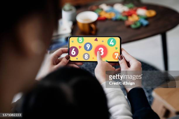 close up of little asian daughter sitting on mother's lap, using smartphone together at home. they are playing educational games on smartphone for e-learning. family lifestyle and technology - child smartphone stock pictures, royalty-free photos & images