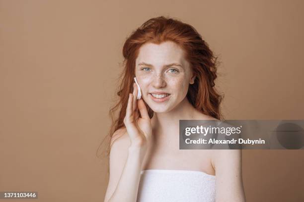 happy redhead woman with freckles takes care of her clean skin by rubbing her face with a sponge on a brown background in the studio. beauty and health concept - máscara facial fotografías e imágenes de stock