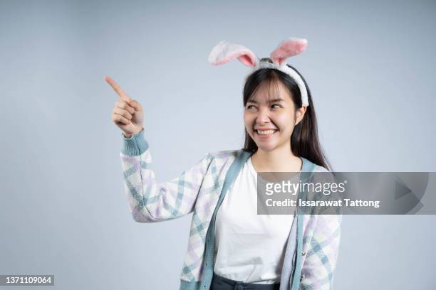 attractive and smiling young girl with bunny ears pointing.picture taken in grey background with copy space. easter holidays. studio shot. - woman fingers in ears fotografías e imágenes de stock