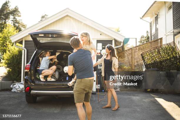 portrait of mother, father and two daughters standing by car at electric vehicle charging station - camino de entrada fotografías e imágenes de stock