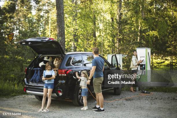 portrait of mother, father and two daughters standing by car at electric vehicle charging station - estación de carga eléctrica fotografías e imágenes de stock