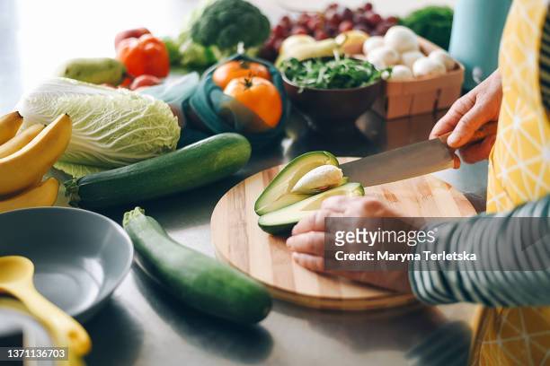 a woman cuts a ripe avocado and takes out a stone from it on a wooden plank. fresh vegetables. salad preparation. diet food. ecological products. - chef table imagens e fotografias de stock