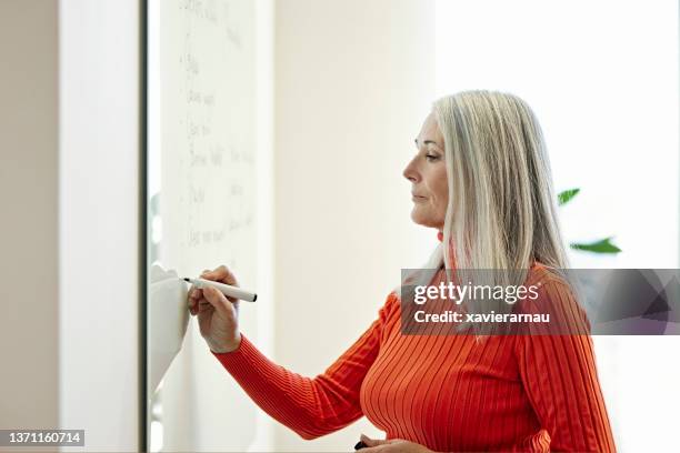 mature businesswoman writing on whiteboard in modern office - female neckline stock pictures, royalty-free photos & images