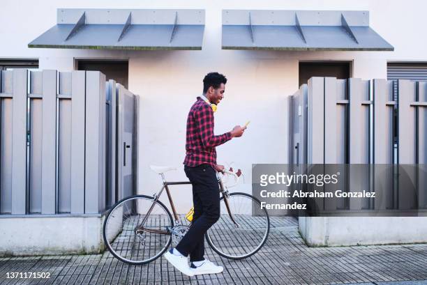 young man walking while pushing his bicycle next to him using his cell phone on sidewalk - casual man walking fotografías e imágenes de stock