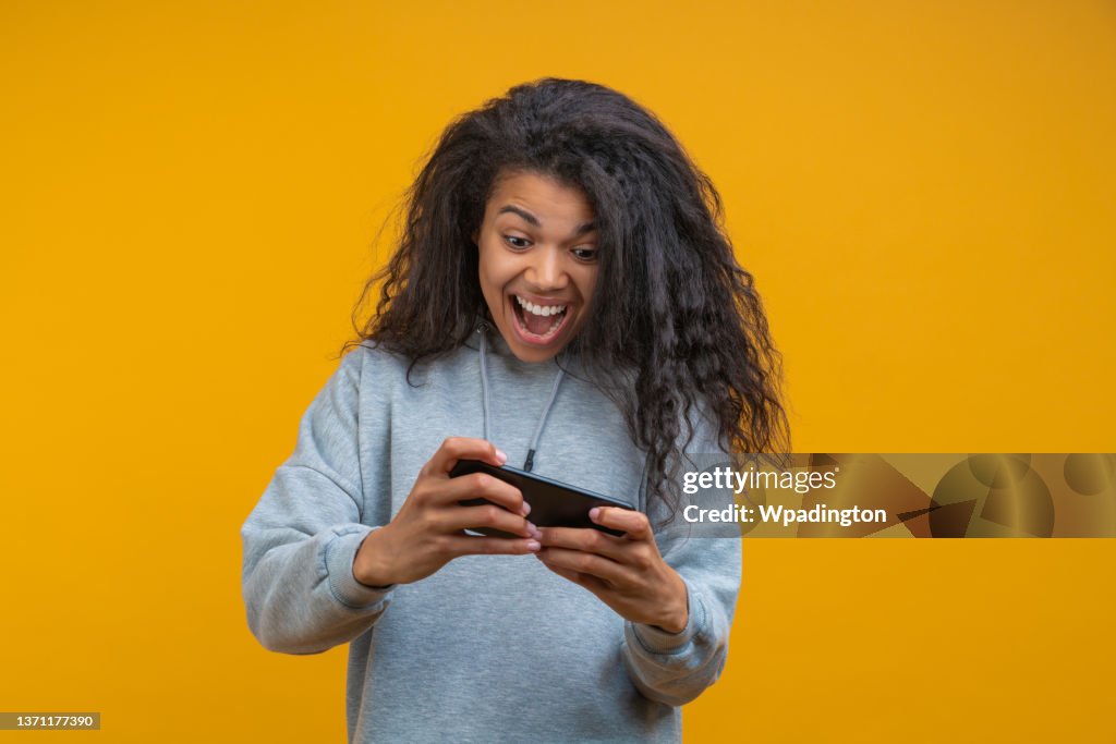 Studio portrait of happy girl reading message with good news on her mobile phone and showing sincere excitement