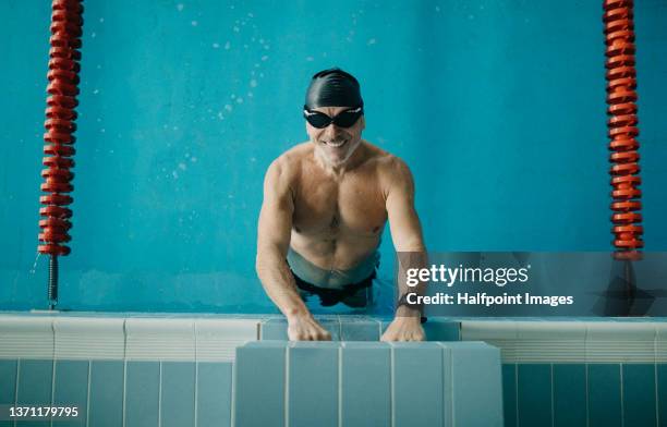 top view of active senior man swimmer holding onto starting block in indoors swimming pool and looking at camera. - swimming stroke stock pictures, royalty-free photos & images