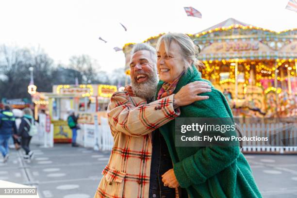 carefree senior couple embracing at outdoor winter carnival - mature couple winter outdoors stock pictures, royalty-free photos & images