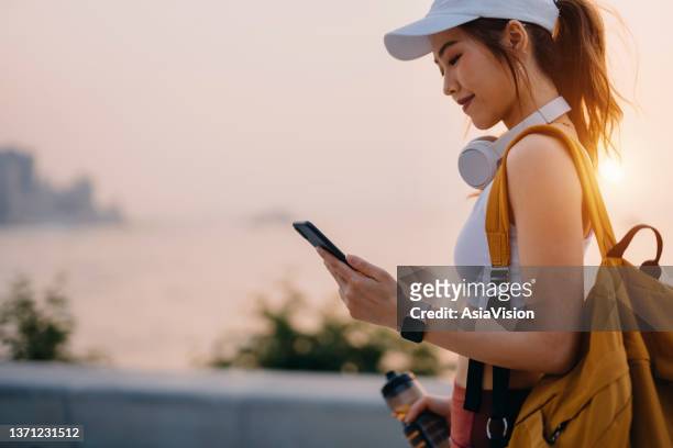 young asian sports woman in sportswear walking along the promenade at sunset, using smartphone after working out in city. she is wearing a cap and headphones, carrying a water bottle and backpack. habits and hobbies. health and fitness with technology - fitnesstracker stock pictures, royalty-free photos & images
