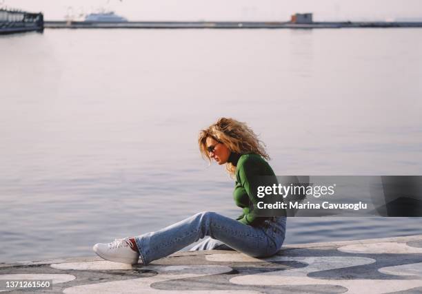 blonde woman sitting at the embankment of a sea making selfie. - frau jeans stock-fotos und bilder