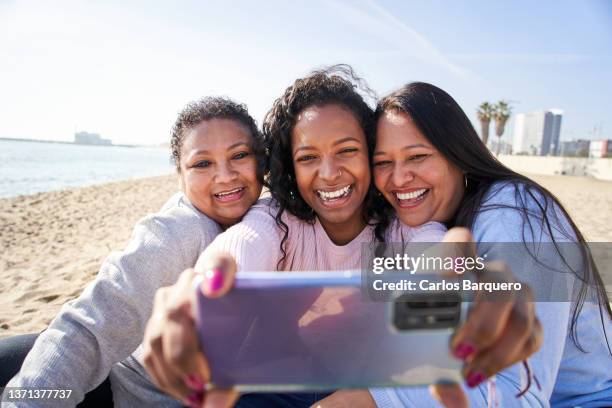 family of three latin american women sitting on the beach and taking a selfie. - mature latin women fotografías e imágenes de stock