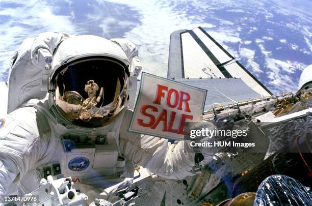 Astronaut Dale A. Gardner, having just completed the major portion of his second extravehicular activity period in three days, holds up a 'For Sale'...