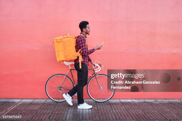 food delivery man with his backpack and bicycle walking along sidewalk - phone side view stock pictures, royalty-free photos & images