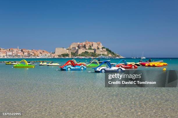 view from beach across clear shallow water to the hilltop citadel, variety of colourful pedalos in foreground, calvi, haute-corse, corsica, france - haute corse stock pictures, royalty-free photos & images