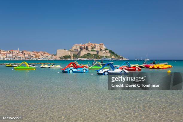 view from beach across clear shallow water to the hilltop citadel, variety of colourful pedalos in foreground, calvi, haute-corse, corsica, france - haute corse bildbanksfoton och bilder