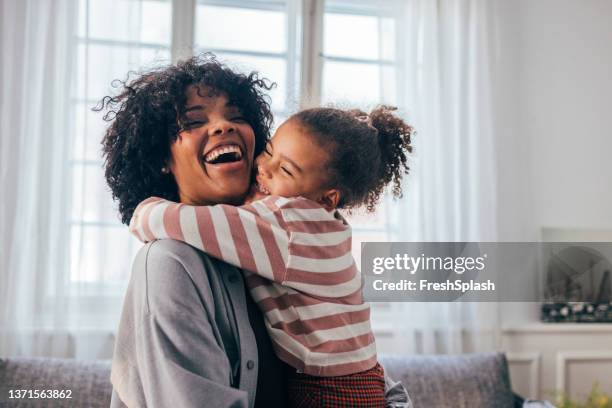 una feliz mujer afroamericana siendo abrazada por su linda hija pequeña en casa - madre fotografías e imágenes de stock