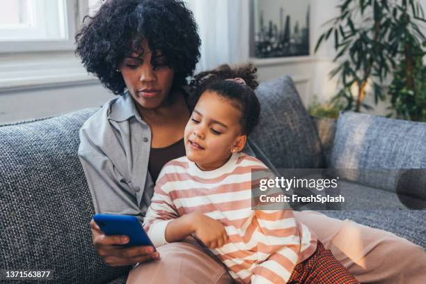 ein entzückendes afroamerikanisches kind, das auf dem sofa sitzt und einen cartoon auf dem handy sieht, das ihre mutter in der hand hält - child animated watching stock-fotos und bilder