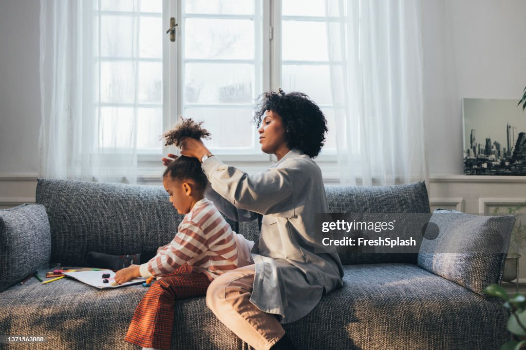 Eine Seitenansicht einer afroamerikanischen Mutter, die die Haare ihrer süßen kleinen Tochter macht, während sie zu Hause auf dem Sofa sitzen