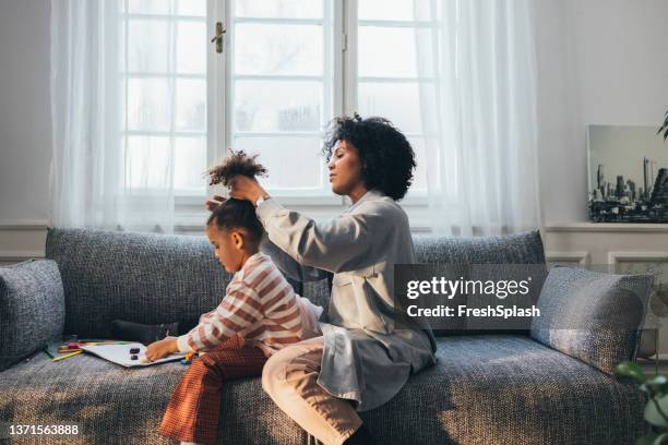 una vista lateral de una madre afroamericana peinando a su linda hija pequeña mientras están sentados en el sofá de casa - fond orange fotografías e imágenes de stock