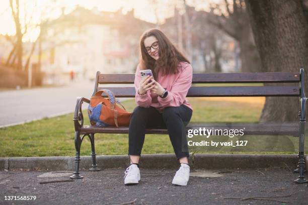 a teenage girl (16-17 years) sitting on a bench in a park texting on her smart phone - 14 15 years stockfoto's en -beelden