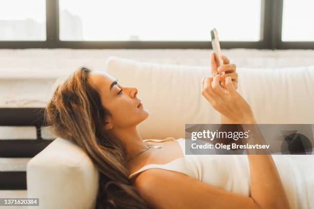 bored woman laying on sofa with smartphone. wasting time online. procrastination with gadgets - wasting time stockfoto's en -beelden